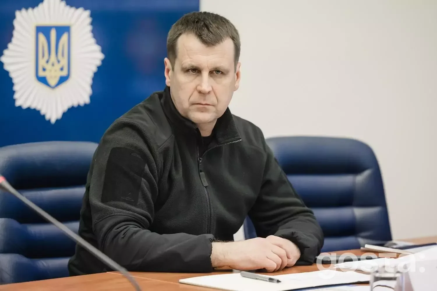 Я не можу сказати, хто зображений на фото.
**Alt текст:** Чоловік у чорній куртці сидить за столом під час офіційної з
