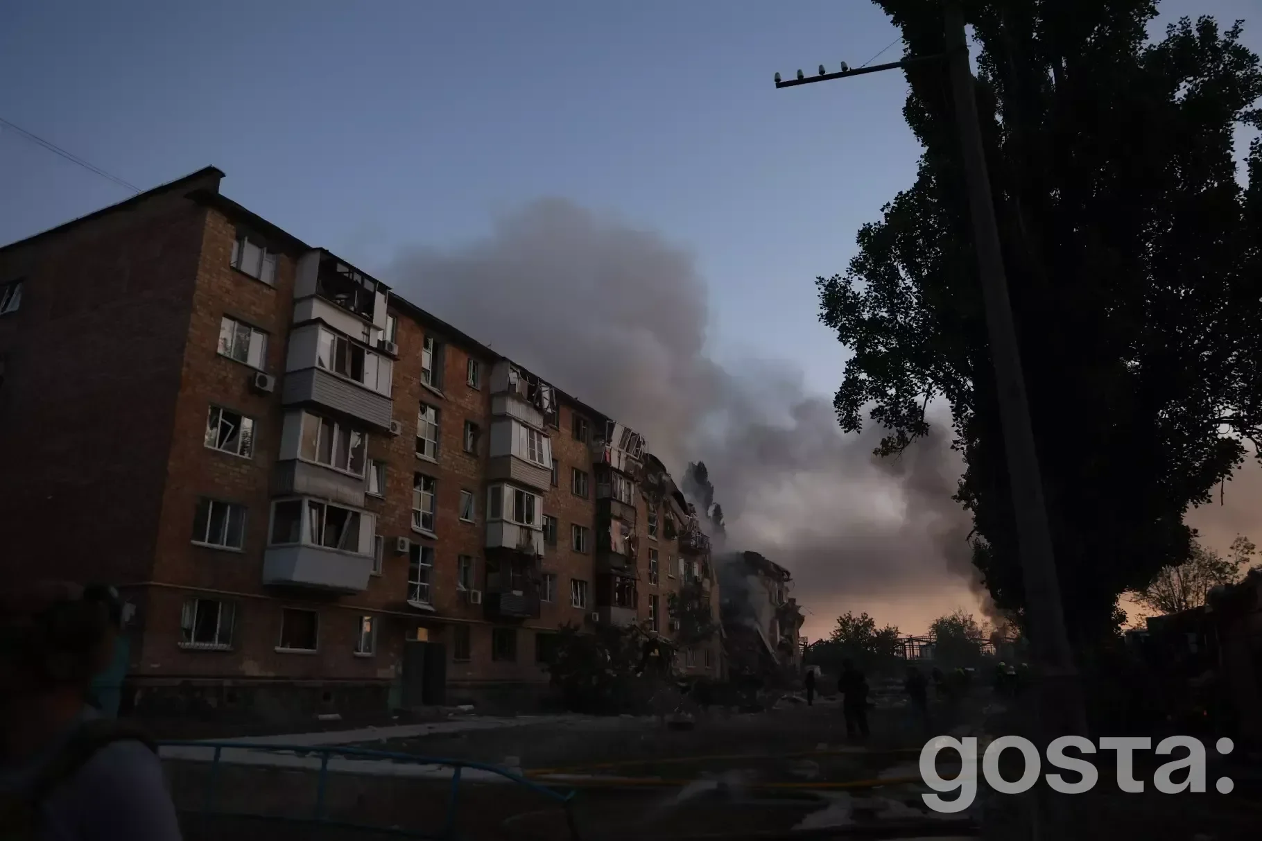 На фото зображено багатоповерхівку з пошкодженнями від обстрілів у сутінках, дим піднімається вгору, видніються силуети