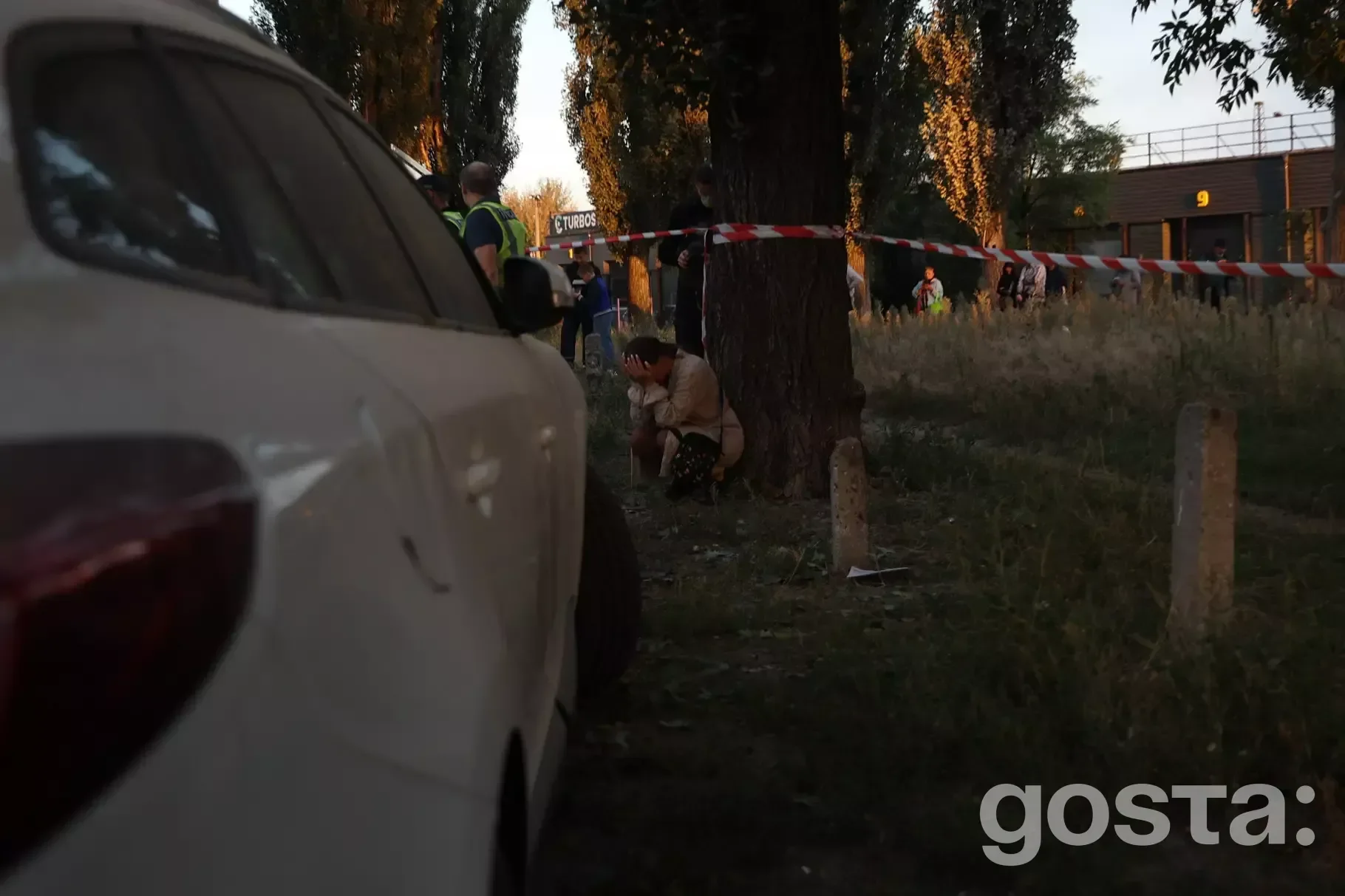 На зображенні бачимо місце події, оточене стрічкою, з поліцейськими і людьми біля дерева у вечірній час.