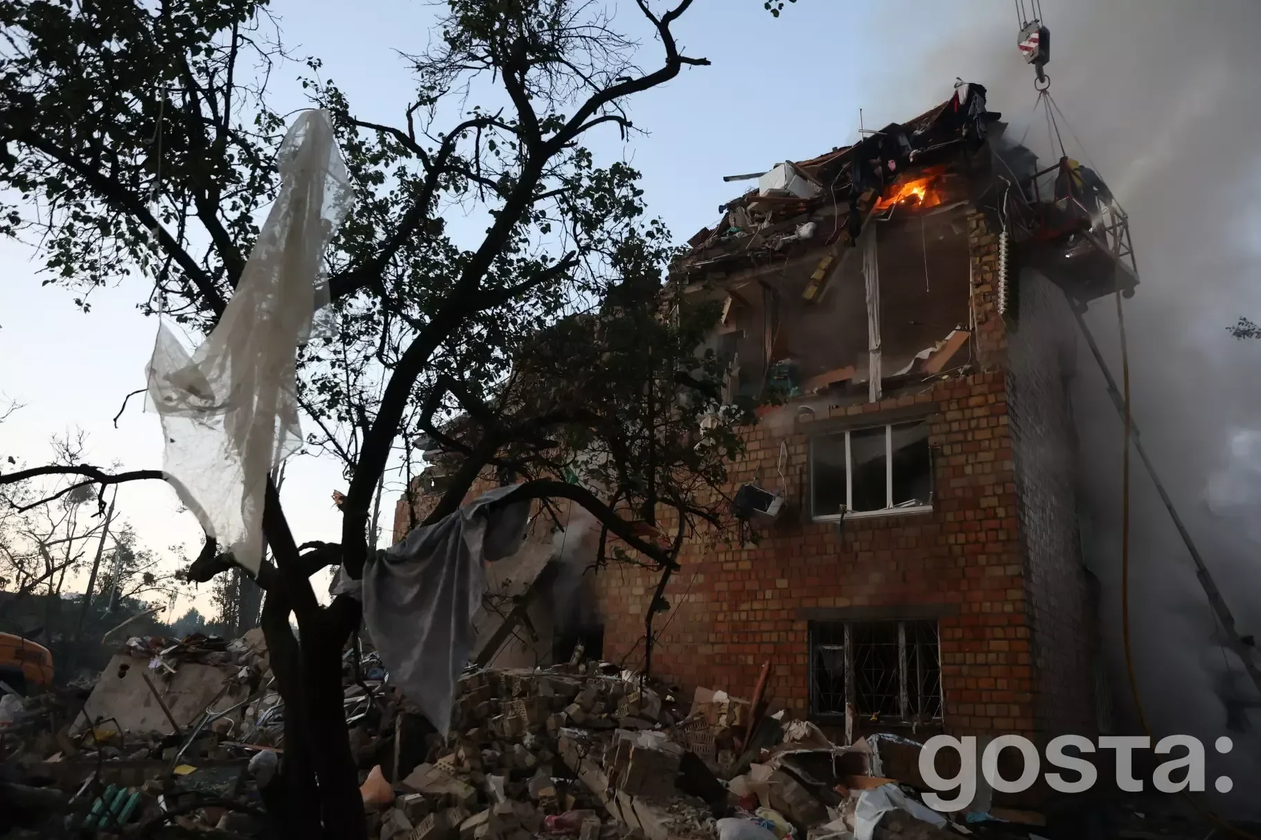 Зруйнований житловий будинок після обстрілу, з пожежою на верхньому поверсі, на тлі вечірнього неба.