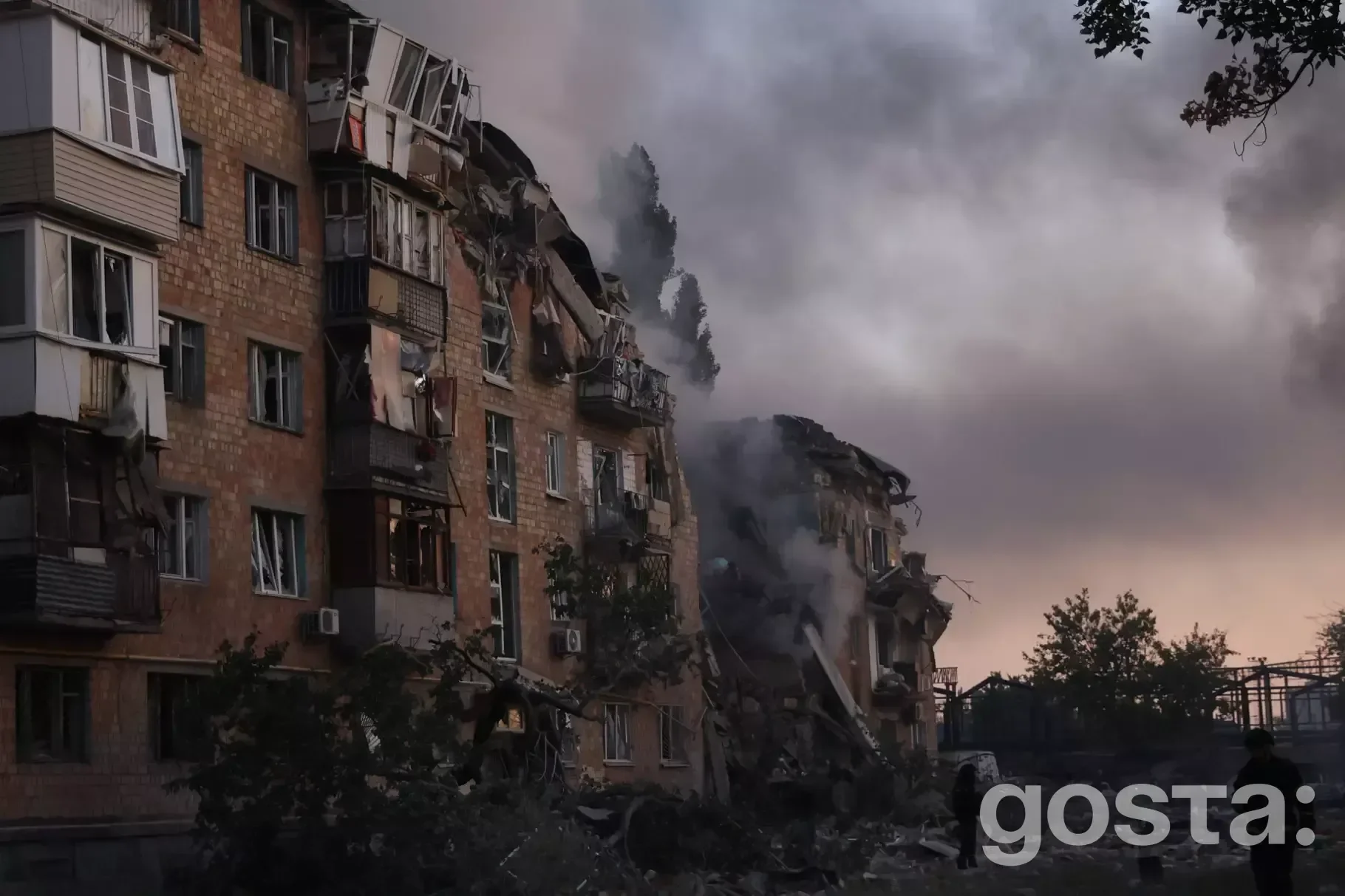 Зруйнована будівля після обстрілу, густий дим піднімається в небо, пошкоджені багатоповерхові будинки.
