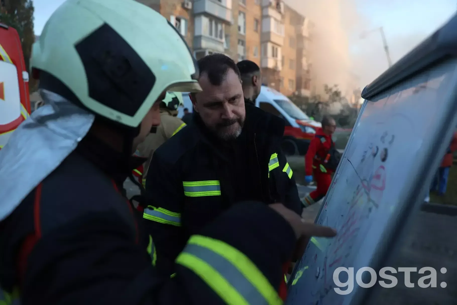 На зображенні пожежники обговорюють план дій біля багатоповерхового будинку з димом на тлі, Україна.