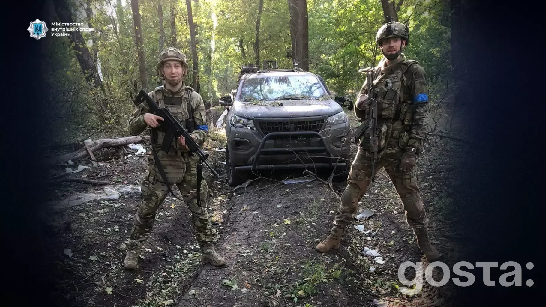 Двоє українських військових стоять у лісі перед позашляховиком, тримаючи зброю, у військовій уніформі.
