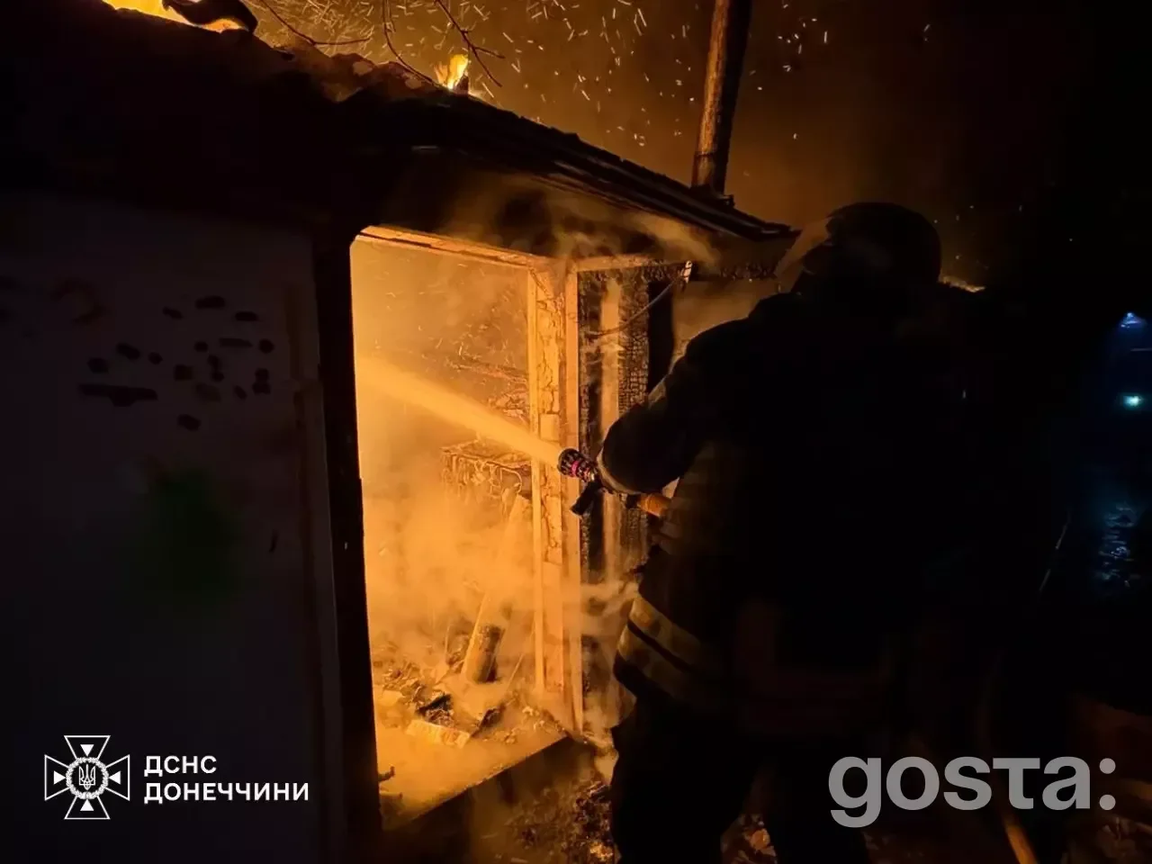 На зображенні зображено пожежника, який гасить палаючу будівлю, іскри та дим навколо, символізує обстріли в Україні.