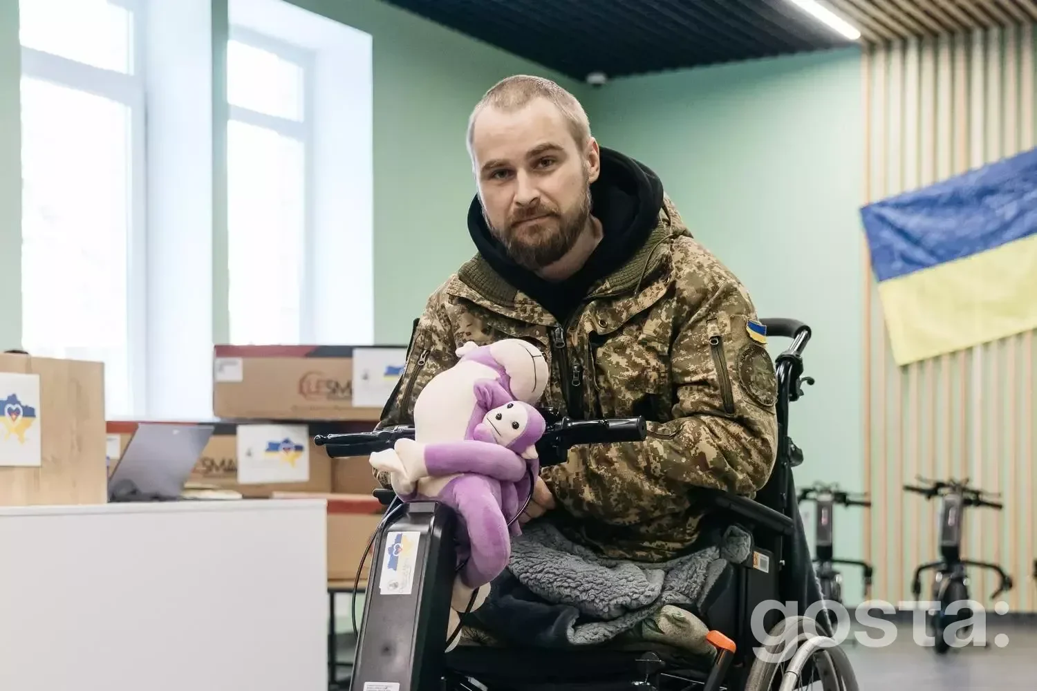 На зображенні: ветеран у камуфляжі сидить у візку з електроприводом, тримаючи м'яку іграшку, у приміщенні медичного закл