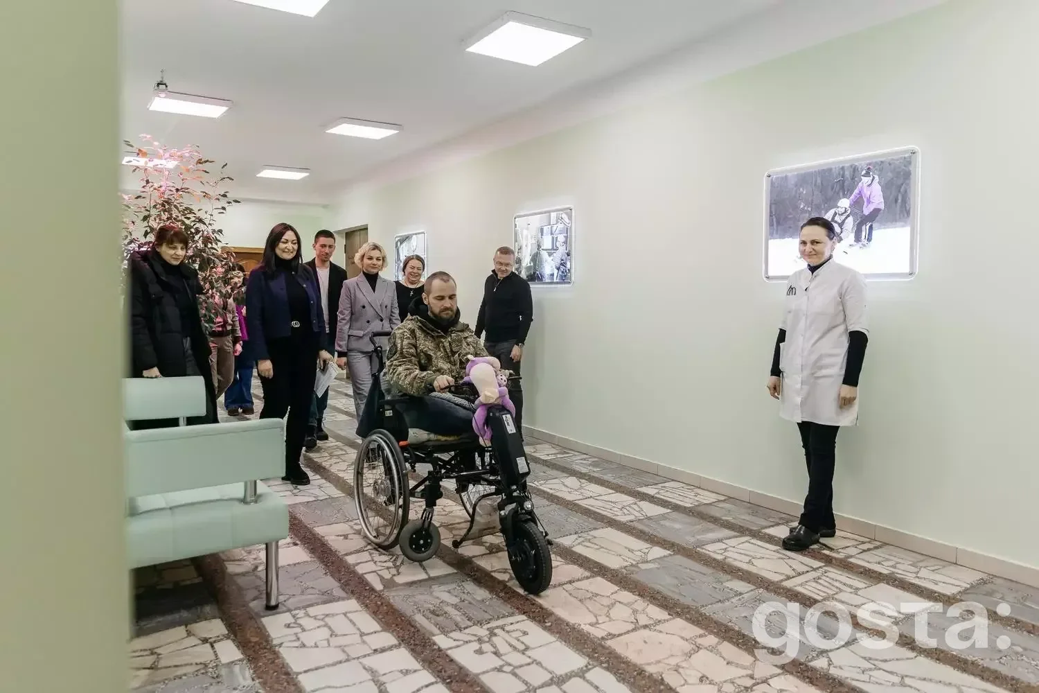 Користувач у кріслі колісному з електроприводом у медичному центрі, оточений медперсоналом і відвідувачами.