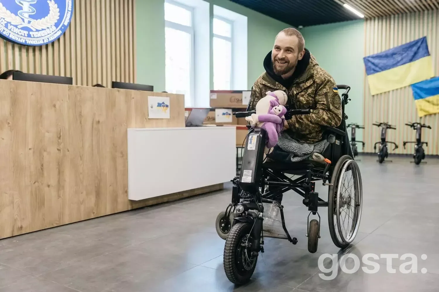 Людина в лікарні їде на електроколясці, посміхаючись, з плюшевою іграшкою на колінах.