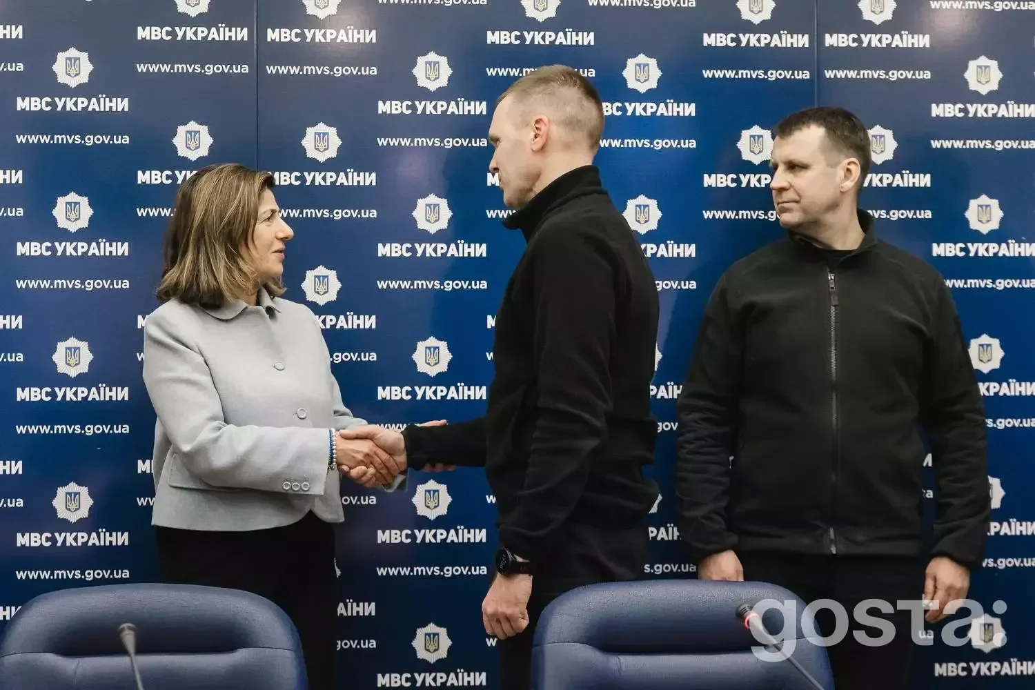 На зображенні представники МВС України і МКЗБ обмінюються рукостисканням на фоні банера "МВС України".