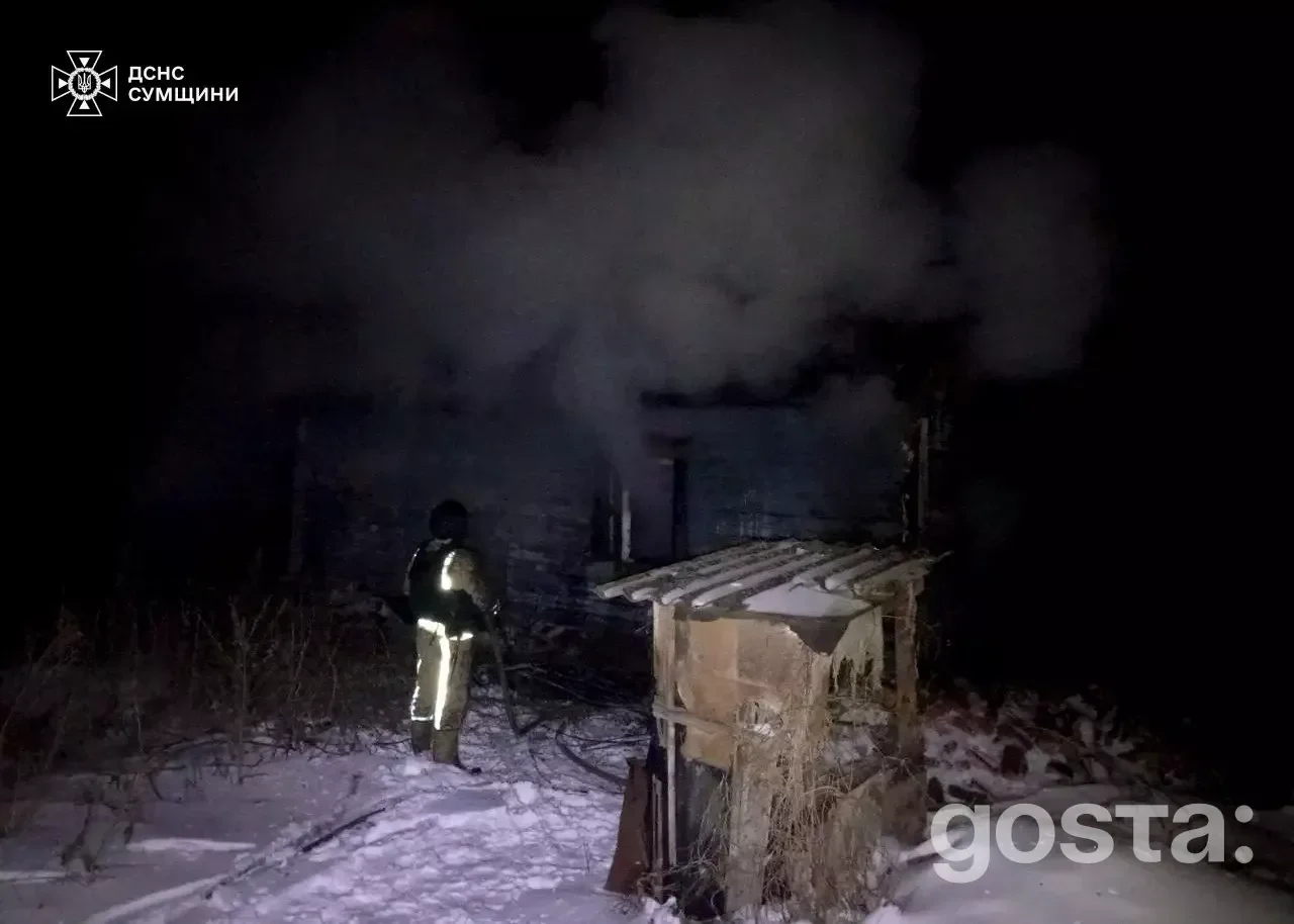На зображенні пожежник гасить палаючу будівлю посеред ночі в засніженій місцевості, дим піднімається в небо.